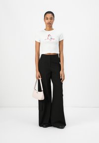 T-shirt blanc court avec un design graphique coloré, associé à un pantalon noir à jambes larges. Tient un petit sac à main rose avec des accents argentés.