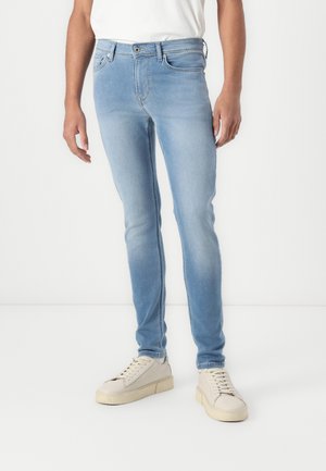 Jean en denim bleu clair avec une coupe slim, présentant un design standard à cinq poches, des passants de ceinture et une fermeture à bouton à l'avant. Associé à des sneakers claires.