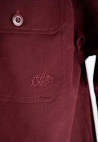 Cartera de camisa burdeos con un solo botón blanco y diseño floral bordado que incluye la letra cursiva "P" en la tela.