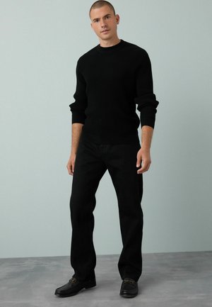 Schwarzer gerippter Strickpullover, schwarze Hose und dunkle Schuhe. Der Schnitt ist lässig, mit einem Rundhalsausschnitt und ärmellangen Ärmeln.