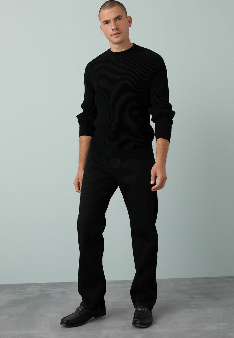 Schwarzer gerippter Strickpullover, schwarze Hose und dunkle Schuhe. Der Schnitt ist lässig, mit einem Rundhalsausschnitt und ärmellangen Ärmeln.