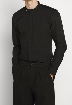 Mand iført en tætsiddende sort mandarin-krave skjorte stukket ned i sorte højtaljede bukser med hænderne i lommerne.