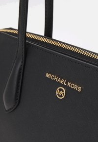 Zwarte handtas gemaakt van gestructureerd materiaal, met een gouden rits, dubbele handvaten en een gouden logo plaat met de tekst "MICHAEL KORS" en een cirkelvormig embleem.