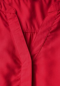 Blouse rouge avec un design en V, ornée de détails froncés au niveau de l'encolure et d'un tissu lisse et léger. Pas de quincaillerie ou de motifs visibles.