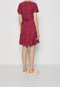 Abito rosso a fiori con maniche corte, orlo svasato e dettaglio in vita con fiocco. Abbinato a scarpe slip-on di colore chiaro e dalla texture particolare.