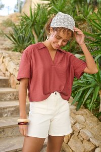 Femme portant un chemisier couleur rouille à col en V, un short blanc, des bracelets décoratifs et un foulard blanc brodé sur la tête, debout à l’extérieur près d’escaliers en pierre.