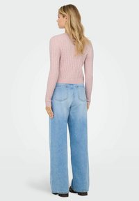 Žena s dugom plavom kosom koja nosi ružičasti džemper od pletene vune i svijetloplave široke traperice, okrenuta leđima prema bijeloj pozadini.