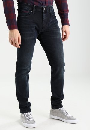 Mężczyzna ubrany w ciemne obcisłe dżinsy, szare wysokie trampki i czerwoną koszulę w granatową kratę na jednolitym tle.