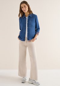 Jeune femme portant une chemise en denim bleue, un haut blanc, un pantalon large beige et des baskets argentées, debout les mains dans les poches.