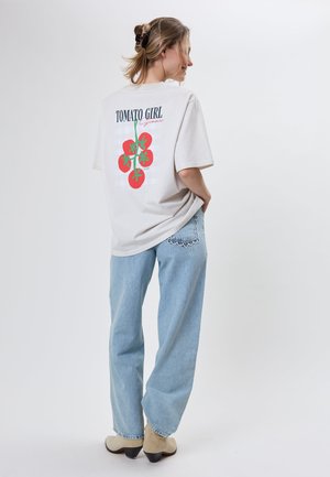 Ung kvinde med samlet hår, iført en hvid T-shirt med røde tomater og teksten "Tomato Girl", lyseblå jeans og beige støvler.