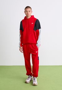 Tânărul stă îmbrăcat într-un trening roșu Nike cu mâneci negre și gri și adidași albi pe un podea verde, în fața unui zid alb.