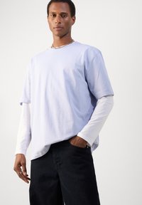 Világos levendula színű, rövid ujjú póló, fehér hosszú ujjú felsőre rétegezve, sötét farmerrel párosítva; az anyag puha és kényelmes, laza szabású.