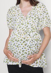 Blouse blanche à motif floral jaune, manches courtes bouffantes, détail noué à la taille, et coupe ample, associée à un bas noir.