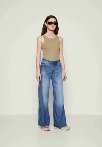 Top de alças bege combinado com jeans largos de lavagens azuis claros, apresentando um acabamento desbotado e bainha desfiada, complementado por calçado bege e óculos de sol pretos.
