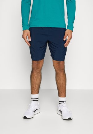 Mężczyzna ubrany w turkusową koszulkę z długim rękawem, granatowe szorty, białe buty sportowe Adidas oraz białe skarpetki z czarnymi paskami, stojący na jednolitym tle.