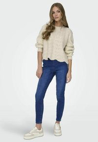 Suéter de punto cable beige claro con un diseño corto, combinado con jeans ajustados azul oscuro y zapatillas color crema con acentos.