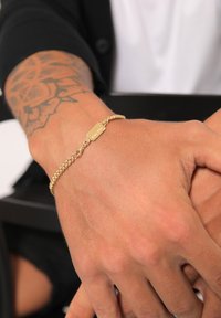 Aukso grandinėlės apyrankė su tekstūruotu dizainu ir stačiakampiu ženklu, išgraviruotu "RUZZO", matoma ant rankos su tatuiruote.