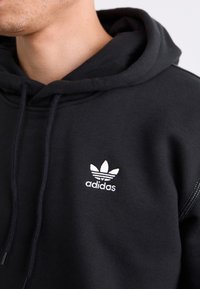 Juodas gobtuvas su kapišonu, pagamintas iš medvilnės mišinio, su baltu Adidas logotipu ir trimis juostelėmis. Apima raištelius kaklo srityje.