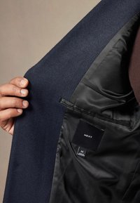 Interior de un blazer de lana azul marino con forro de satén negro, que presenta un bolsillo y una etiqueta que muestra la marca "NEXT" y la talla "M".