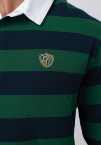 Ράγκμπι πουκάμισο με οριζόντιες πράσινες και σκούρες μπλε ρίγες, λευκό γιακά και κεντημένο έμβλημα ασπίδας στο στήθος.