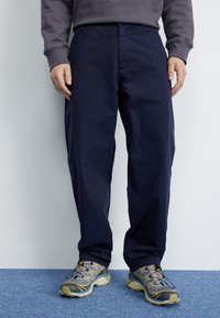 Personne portant un pantalon large bleu foncé, des chaussures de sport grises avec accents noirs et jaunes, se tenant sur un tapis bleu contre un mur blanc.