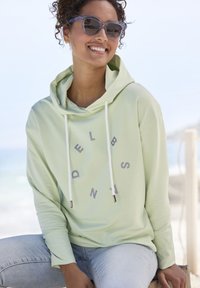 Felpa con cappuccio verde chiaro con scritte grigie, dotata di cordini bianchi e punte in tonalità argento, indossata con jeans azzurri chiari.