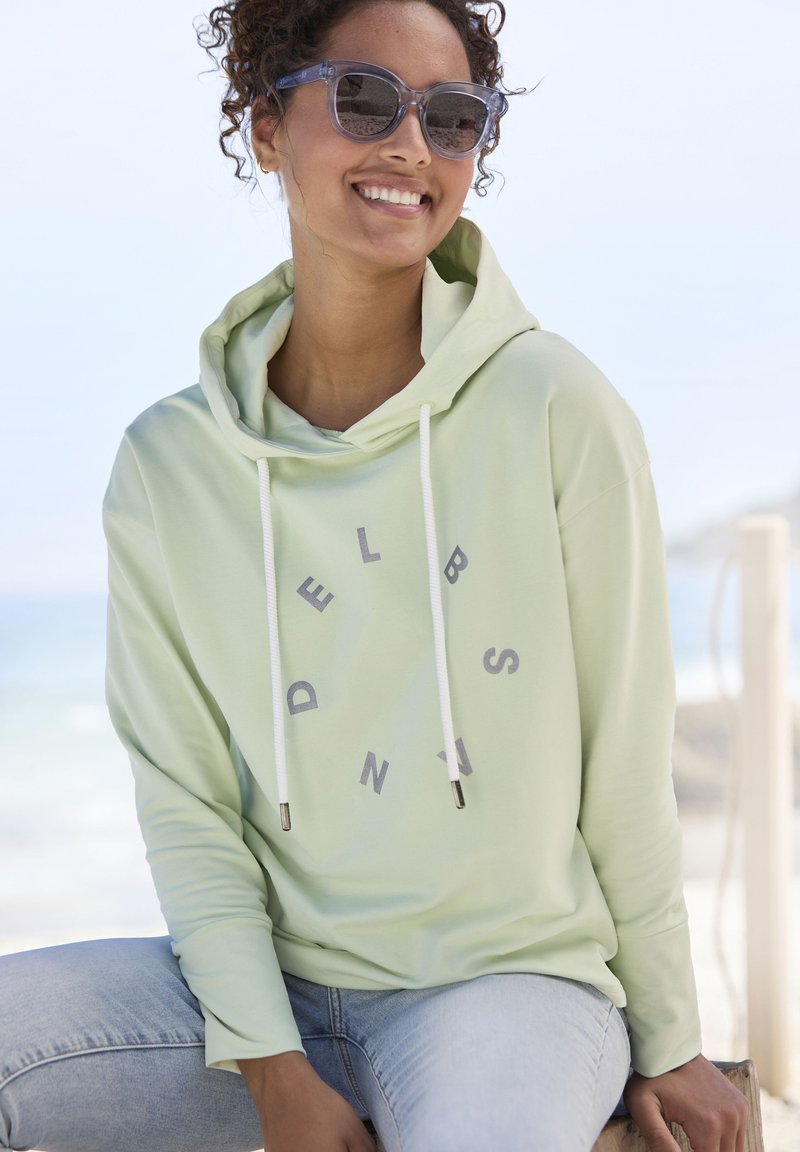 Felpa con cappuccio verde chiaro con scritte grigie, dotata di cordini bianchi e punte in tonalità argento, indossata con jeans azzurri chiari.