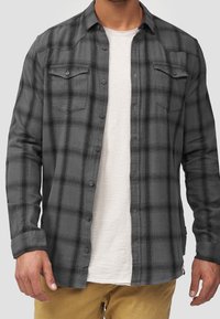 Chemise en flanelle à carreaux gris et noir, dotée de manches longues, deux poches poitrine, fermeture par boutons, et une coupe décontractée portée sur un t-shirt clair.