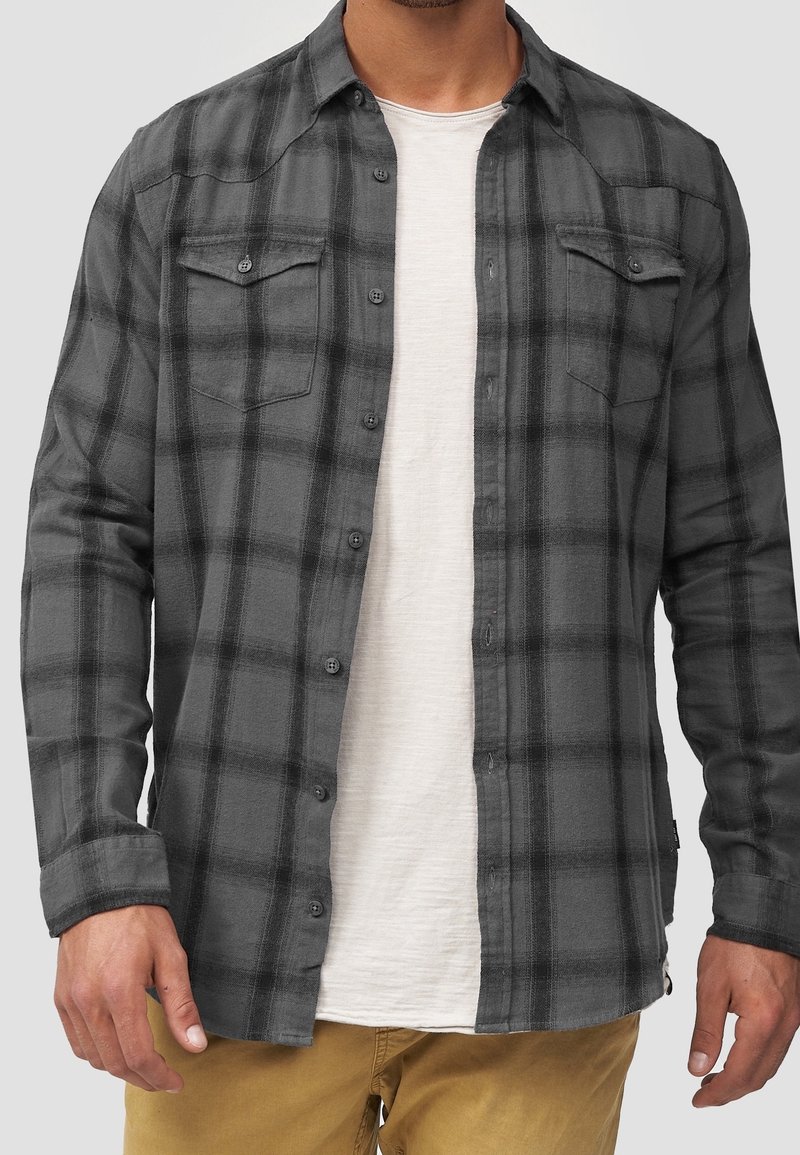 Chemise en flanelle à carreaux gris et noir, dotée de manches longues, deux poches poitrine, fermeture par boutons, et une coupe décontractée portée sur un t-shirt clair.