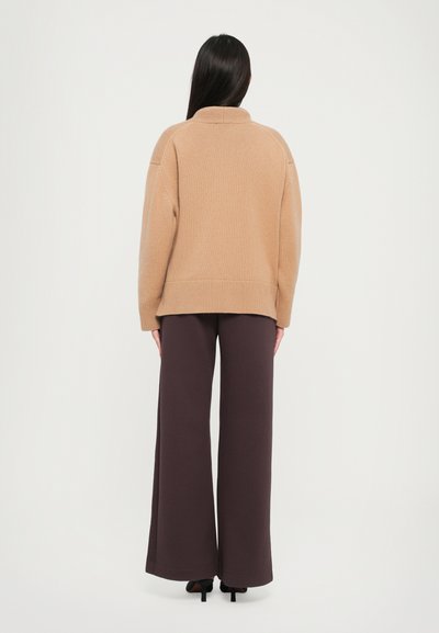 Beige oversize pulóver magas gallérral, bordázott mandzsettákkal és aljjal, sötétbarna, bő szárú nadrággal és fekete magassarkú cipővel párosítva.