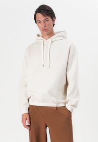 Crème kleurige hoodie van zachte stof met trekkoorden, elastische zoom en subtiele logo-details. Gecombineerd met bruine slim-fit broek.