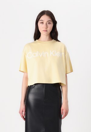 Geel cropped t-shirt met korte mouwen, voorzien van witte "Calvin Klein" logo, gecombineerd met een zwarte leren rok. Soepele textuur.