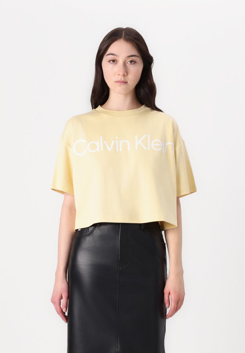 Gul croppet t-skjorte med korte ermer som har hvit "Calvin Klein" logo, kombinert med et svart skinnskjørt. Glatt tekstur.