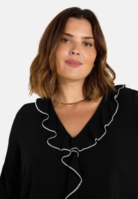 Blusa nera con scollatura a V e un bordo arricciato femminile con filo metallico. Tessuto morbido, vestibilità comoda e maniche corte. Accento di collana dorata.