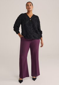 Blusa nera texture con scollo a V e bottoni frontali, abbinata a pantaloni larghi viola a vita alta e décolleté neri a punta.
