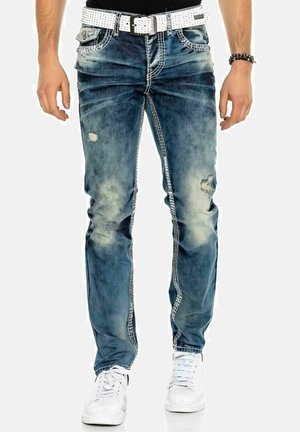 Hombre con vaqueros azules desgastados y desteñidos con costuras blancas, un cinturón blanco perforado, pulseras negras con cuentas y zapatillas blancas.