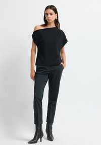 Top nero scollato a spalla con vestibilità ampia, abbinato a pantaloni verdi scuri su misura e stivali neri con tacco a punta, su uno sfondo neutro.