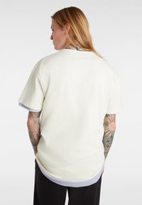 Camiseta color crema con un ajuste holgado, mangas cortas y dobladillo en capas. La tela parece suave con una textura lisa; detalles incluyen costuras visibles.