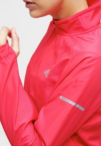 Primer plano de una persona vestida con una chaqueta de running Adidas de un rosa brillante, con media cremallera y una franja reflectante "adidas running" en la manga.
