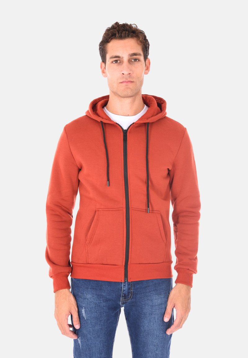 Felpa con zip color ruggine, con tasca frontale, lacci neri e polsini a costine, indossata sopra una maglietta bianca, abbinata a jeans blu.