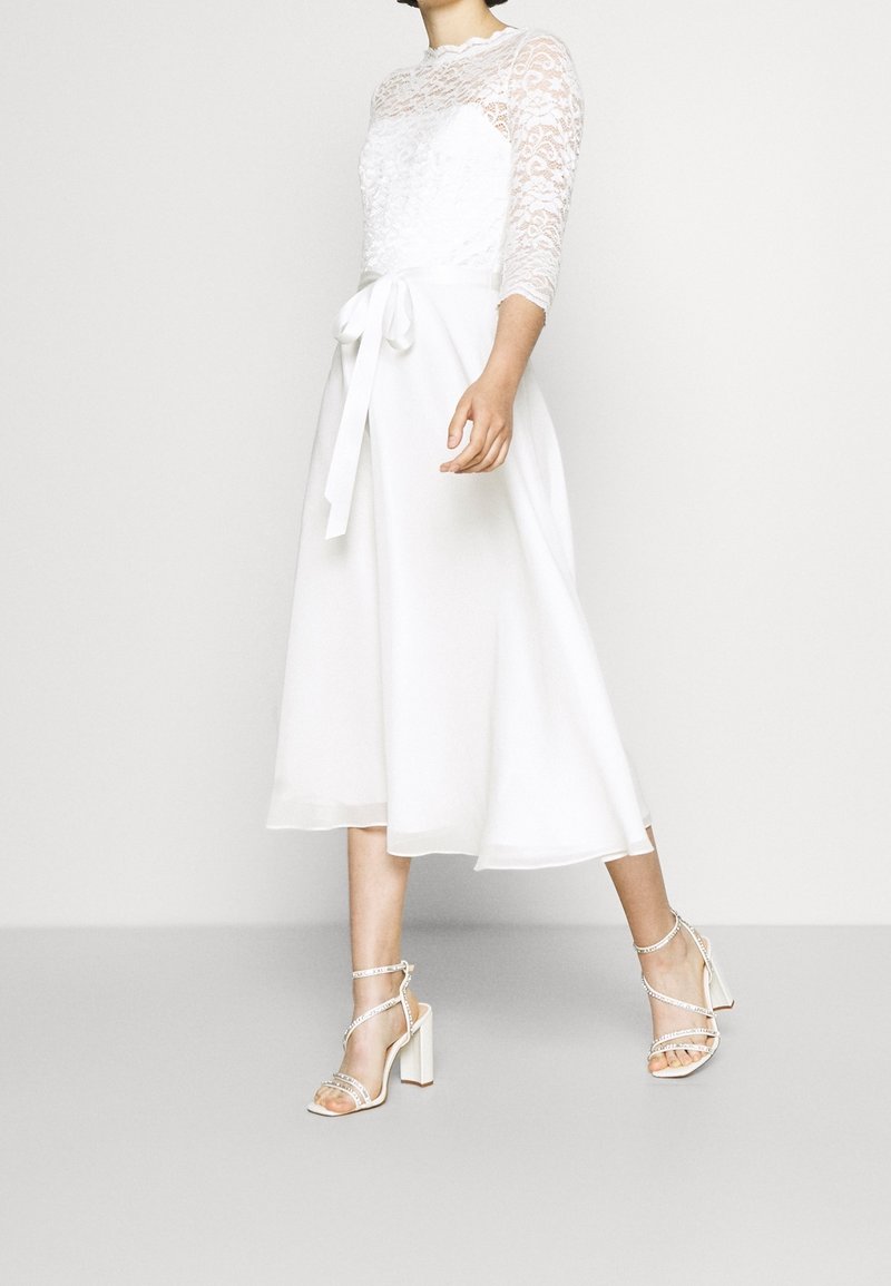 Femme portant une robe midi en dentelle blanche avec une ceinture nouée à la taille et des sandales blanches à talons hauts et open-toe, debout devant un fond uni.