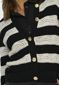 Cardigan rayé noir et blanc avec texture en crochet et boutons dorés, comportant des sections alternées unies et à motifs.