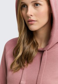 Sudadera rosa con un tejido suave y texturizado, cierre con cordón y capucha, con detalles acanalados visibles en los hombros y las mangas.