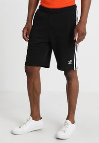 Shorts neri in cotone con strisce bianche Adidas sui lati, dotati di una cintura elastica e tasche laterali. Abbinati a scarpe bianche.
