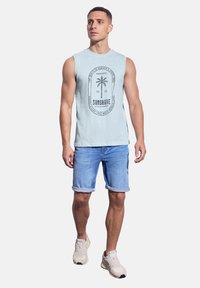 Svetle modré tílko bez rukávů s grafickým motivem palmy a textem, v kombinaci s ohrnutými džínovými šortkami. Modelka má na sobě béžové tenisky.