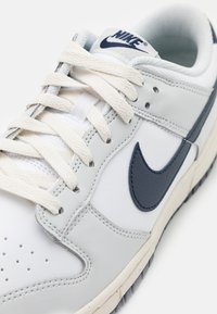 Sneaker bianca in pelle con accenti blu navy, dotata di punta rotonda, perforazioni e lacci beige. Include un logo Nike testurizzato blu.