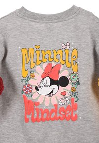 Grauer Pullover mit einem bunten Motiv eines lächelnden Charakters mit roter Schleife, umgeben von Blumen und dem Schriftzug "Minnie Mindset".