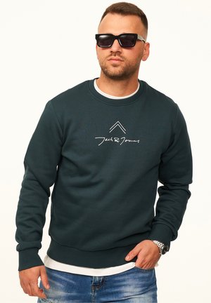 Mann mit schwarzen Sonnenbrillen, dunkelgrünem Jack & Jones Sweatshirt, weißem Unterhemd, blauen Jeans und silberner Armbanduhr, der vor weißem Hintergrund posiert.