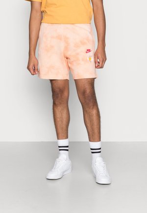 Nike-shorts in licht perzik tie-dye, met een geborduurde zon en logo. Gecombineerd met witte sneakers en zwart-gestreepte sokken.