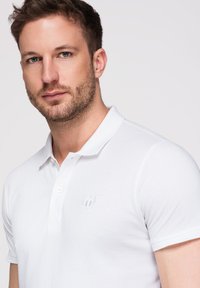 Homme aux cheveux bruns courts et à la barbe portant un polo blanc à manches courtes sur un fond clair.